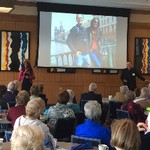 A woman and a man stand and speak in front of a screen that shows a picture of the two of them in Russia, while 20 seated people are listening